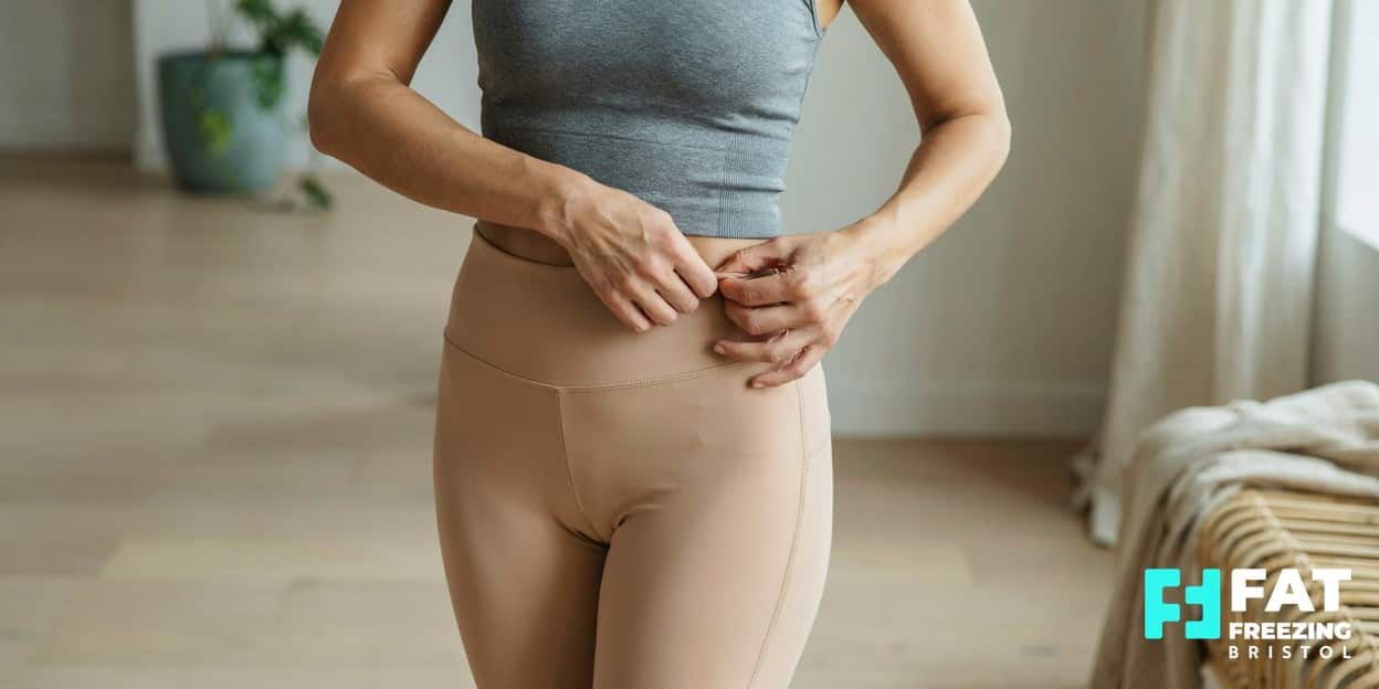 A person in activewear gently pinching a small pocket of skin at the lower abdomen in a pinch-test gesture, illustrating why stubborn fat pockets can resist diet and exercise alone