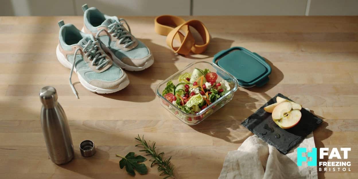 A still-life of running trainers beside a container of fresh salad, a sliced apple, a water bottle and a resistance band on a sunlit kitchen counter, representing the healthy lifestyle that works alongside fat freezing